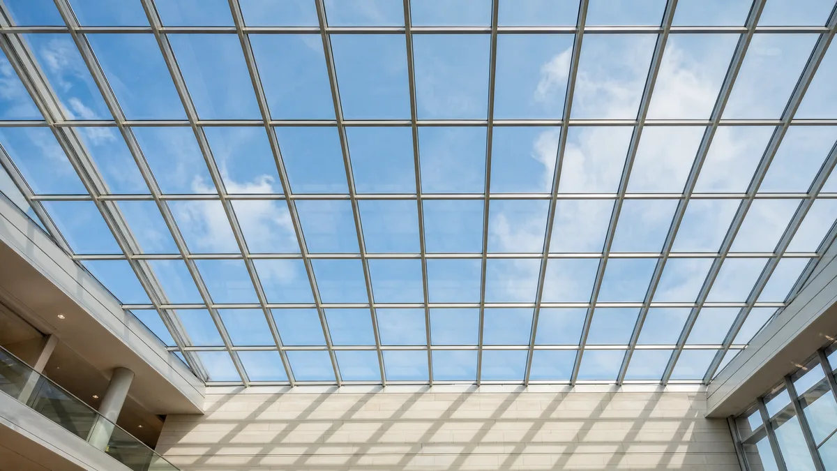 Blick von unten auf ein Glasdach über einem Atrium: Sprossenraster vor blauem Himmel, Gitterschatten auf der Innenwand